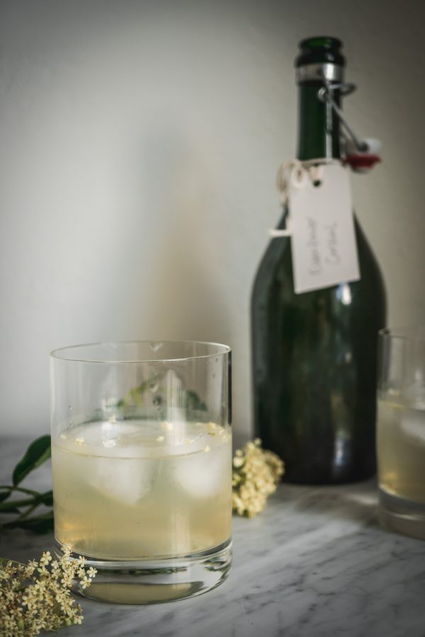 Cocktail glass filled with elderflower cordial and ice cubes sitting on a marble counter.