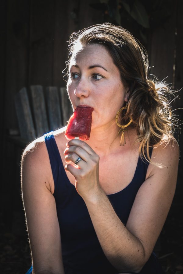 Woman in a blue dress sits outside and eats a strawberry popsicle. 