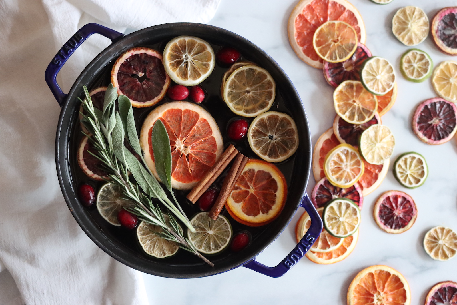 Simmer pot with dried citrus slices, fresh cranberries, cinnamon sticks, rosemary, and sage in a blue braiser